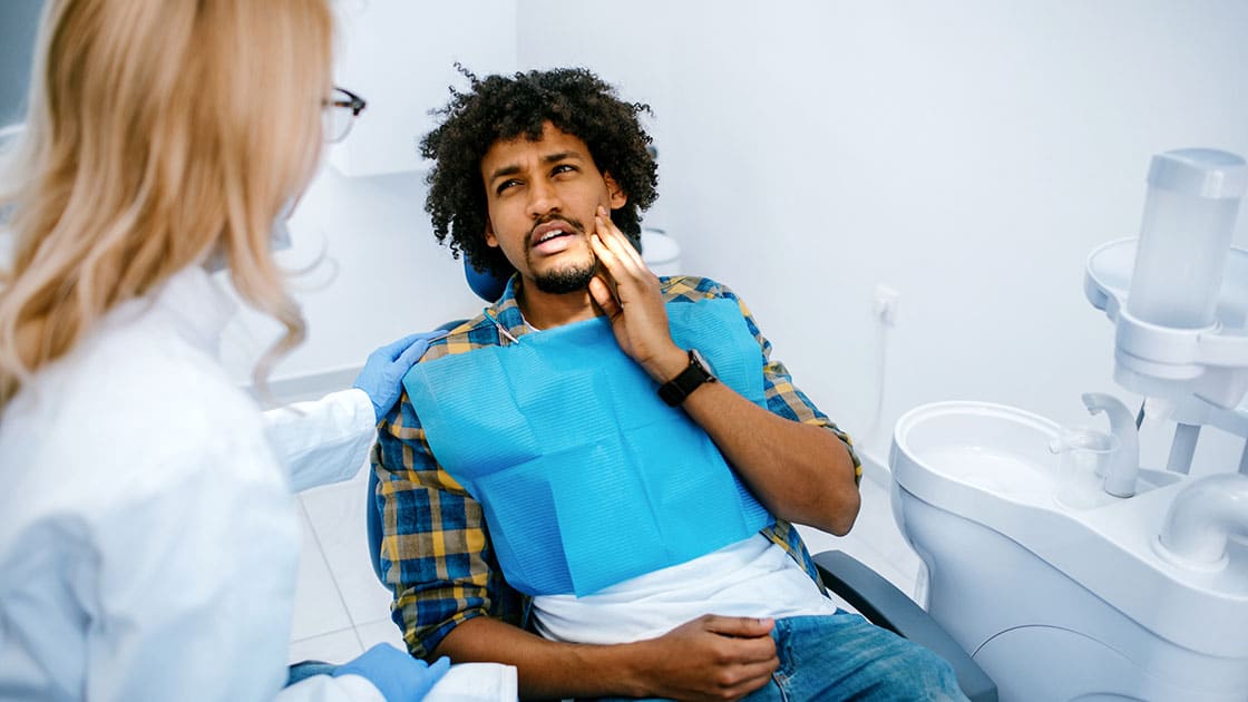 Young black man holding cheek in chair at dentist, having toothache.